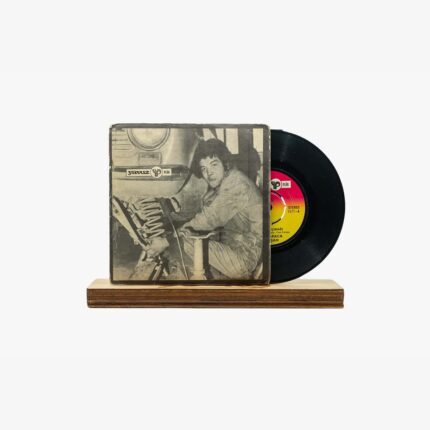 Vintage vinyl record partially pulled from a worn sleeve, resting on a wooden stand, with a black disc and a grayscale photo on the cover showing a person in a studio.