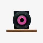 2301450079C | 3345 Records Black vinyl record mounted on a dark square display block, with a pink center label, set on a wooden base.