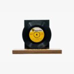 2301450095B | 3345 Records Black vinyl record mounted on a wooden display stand with a yellow label at its center, against a white background.